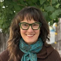 Gina di Grazia — Orientation and Mobility Specialist & Low Vision Therapist Smiling woman with wavy dark hair wearing green glasses and a blue scarf, standing outdoors near trees.