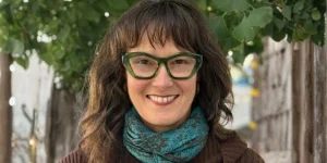 Smiling woman with wavy dark hair wearing green glasses and a blue scarf, standing outdoors near trees.