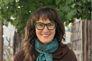 Smiling woman with wavy dark hair wearing green glasses and a blue scarf, standing outdoors near trees.