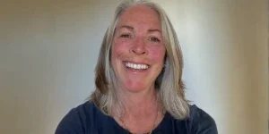 Smiling woman with shoulder-length gray hair wearing a black top, standing indoors against a light background.