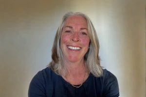 Smiling woman with shoulder-length gray hair wearing a black top, standing indoors against a light background.