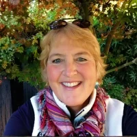 Kathy Abrahamson — Rehabilitation Teacher Smiling woman with blonde hair wearing a scarf and dark jacket, standing outdoors with greenery in the background.