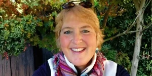 Smiling woman with blonde hair wearing a scarf and dark jacket, standing outdoors with greenery in the background.