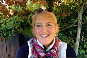 Smiling woman with blonde hair wearing a scarf and dark jacket, standing outdoors with greenery in the background.