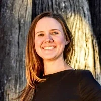 Kelly Whittlesey — Ergonomics Specialist Smiling woman with straight light-brown hair wearing a black top, standing outdoors near a tree.