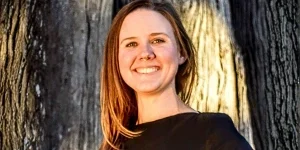 Smiling woman with straight light-brown hair wearing a black top, standing outdoors near a tree.