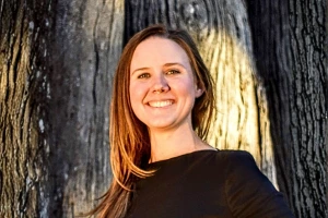 Smiling woman with straight light-brown hair wearing a black top, standing outdoors near a tree.