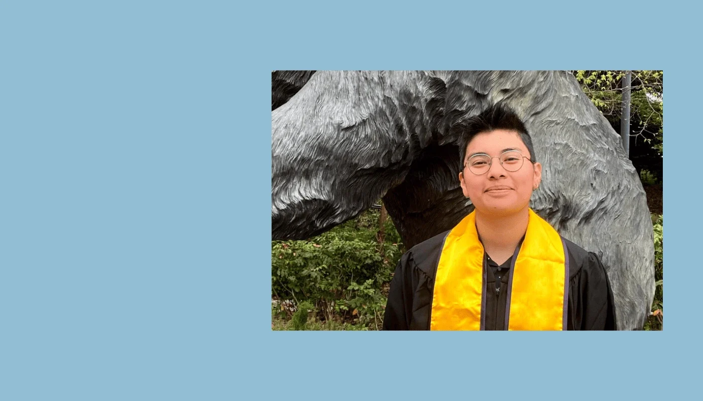 Smiling person with short hair and glasses wearing a graduation gown with a yellow stole, standing outdoors near a large sculpture.