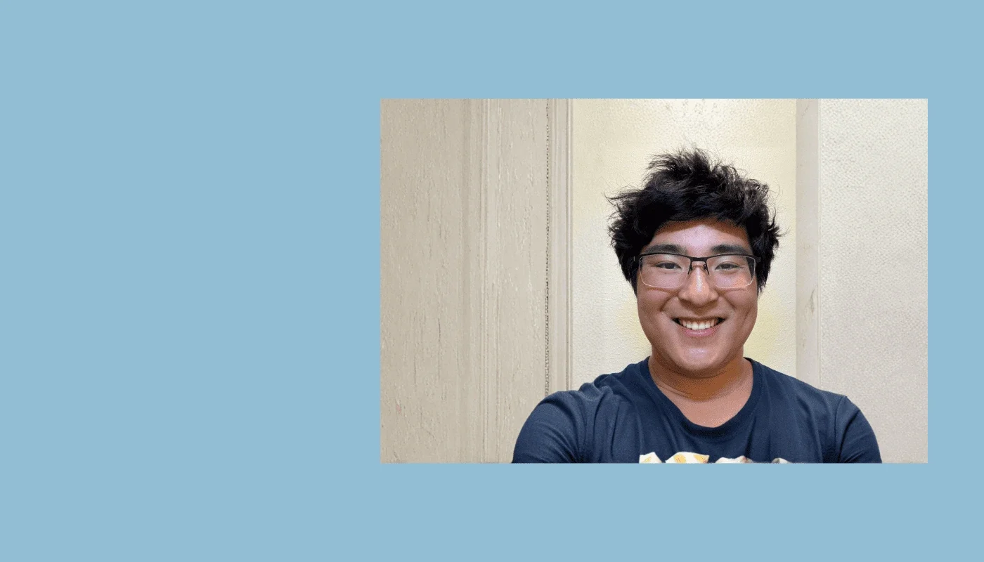 Smiling young man with glasses wearing a navy blue shirt, standing indoors against a light-colored wall.