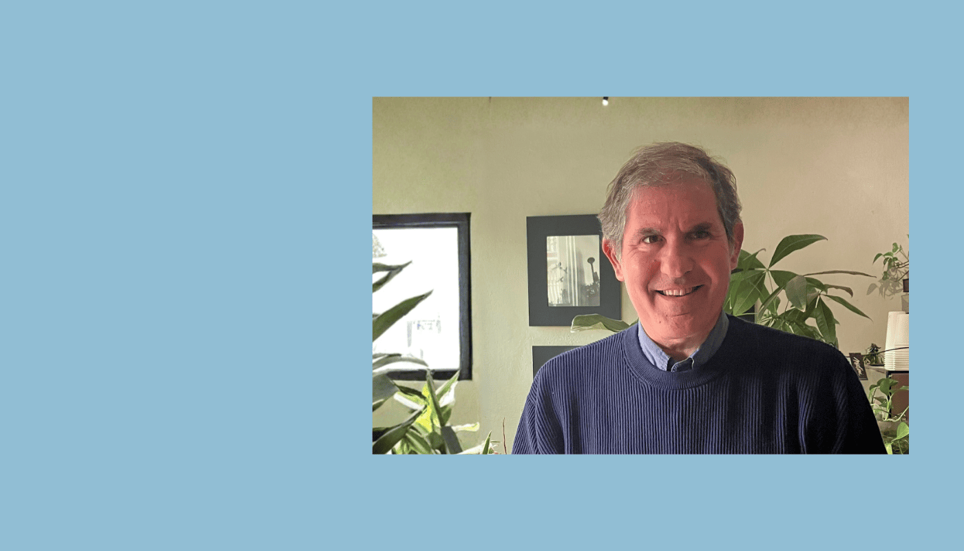 Smiling man with gray hair wearing a navy blue sweater, standing indoors with green plants in the background.