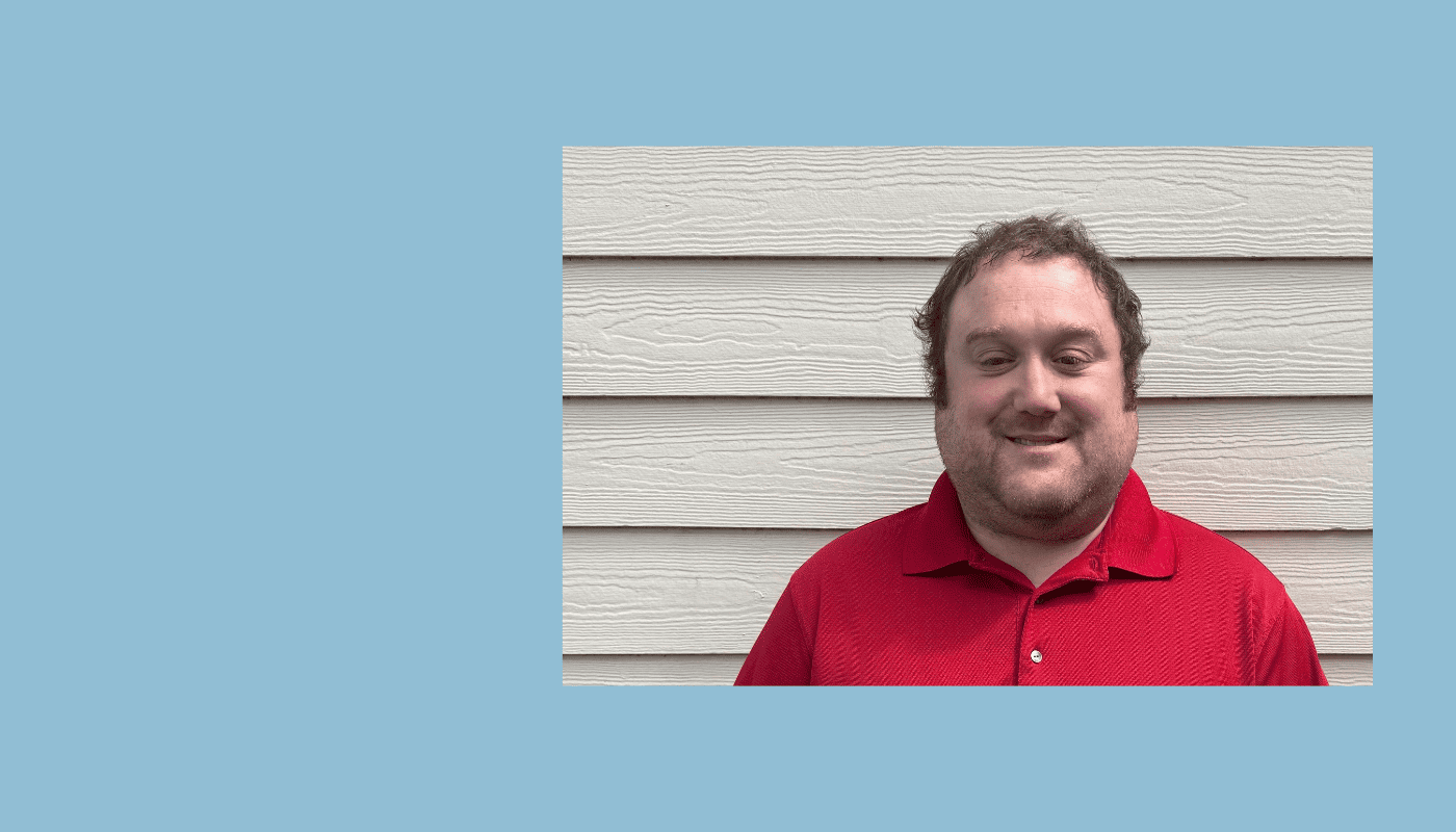Smiling man wearing a red collared shirt standing in front of a light-colored wall.