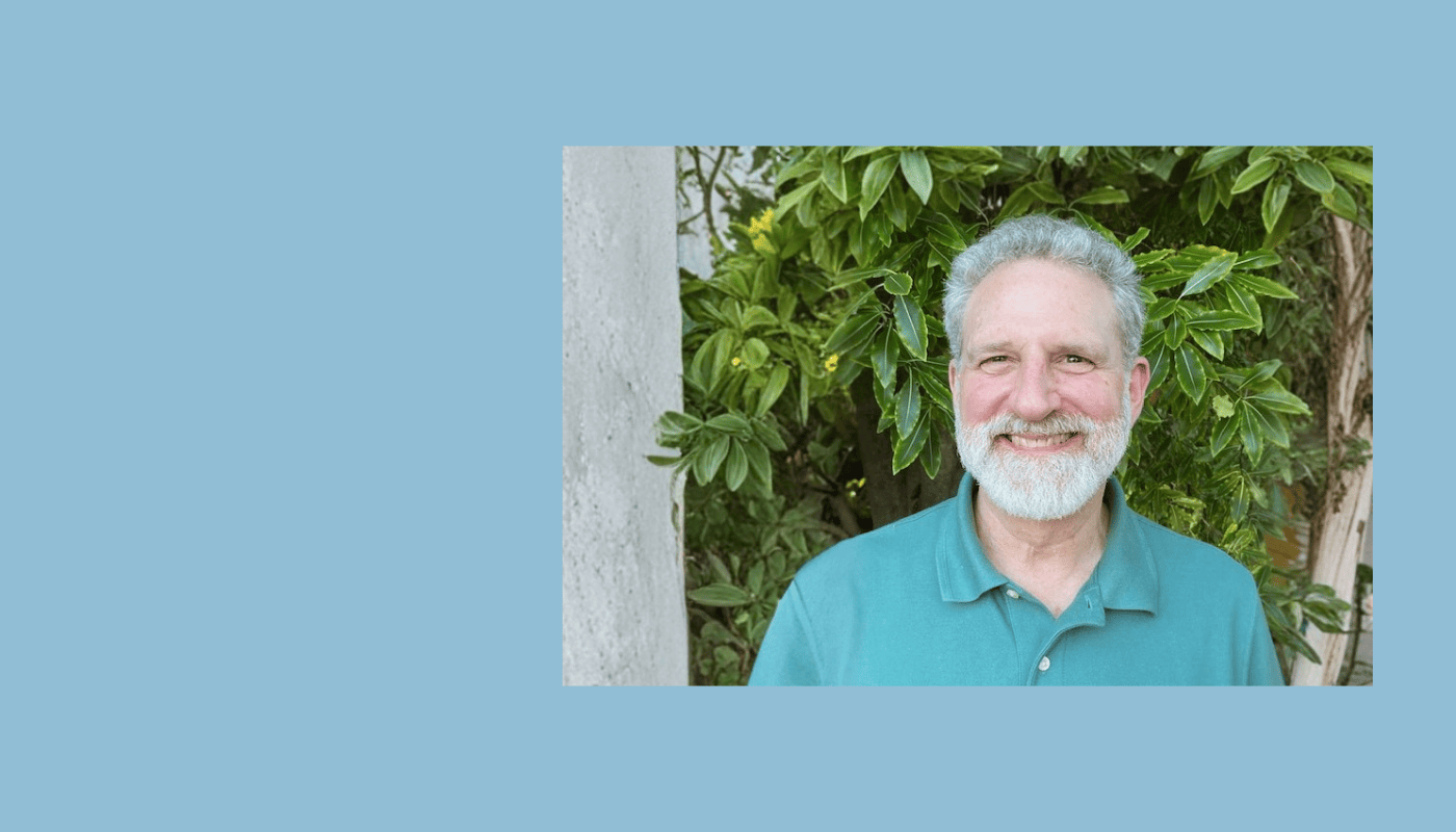 Smiling man with gray hair and beard wearing a teal shirt, standing in front of green foliage outdoors.