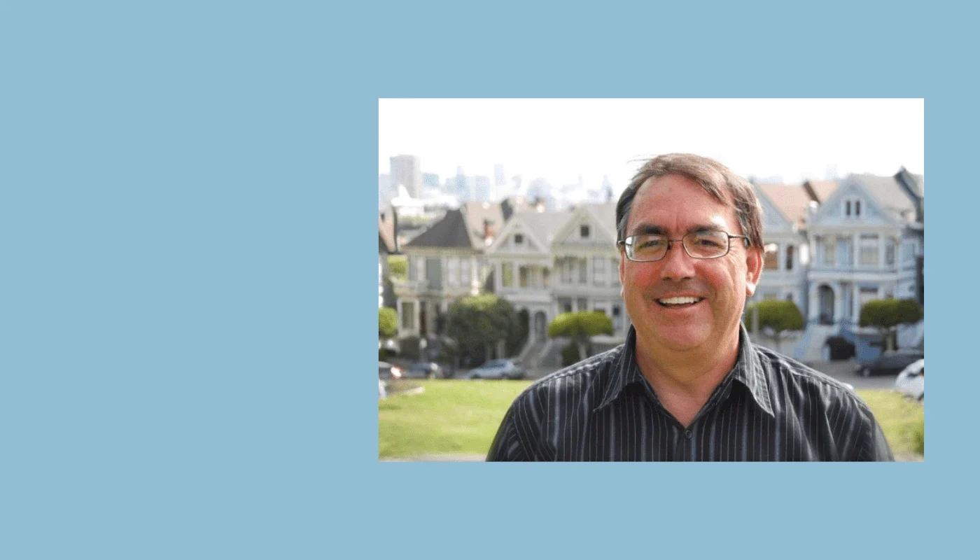 Smiling man with glasses standing outdoors with Victorian-style houses in the background, representing the co-founder of Adaptive Technology Services.
